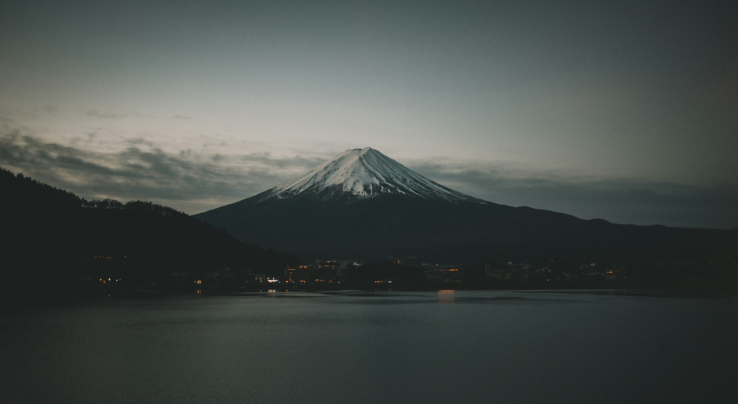 Dusk over a still lake with a snow-capped volcano beyond and the lights of a small town along the shore.